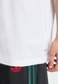Λευκό βαμβακερό T-shirt με μικρό μαύρο λογότυπο Adidas σε ταινία στο πλάι. Φορεμένο με σκούρο γκρι παντελόνι που φέρει κόκκινο λογότυπο και πράσινες ρίγες.