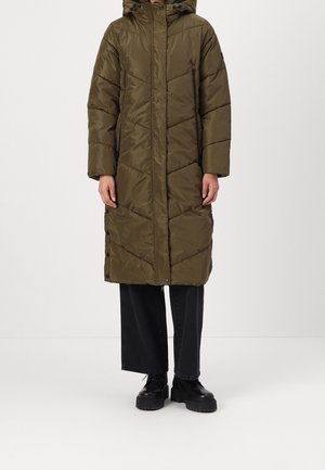 Personne portant un long manteau matelassé vert olive avec une capuche, un pantalon noir à jambes larges et des chaussures noires épaisses, debout devant un fond uni.