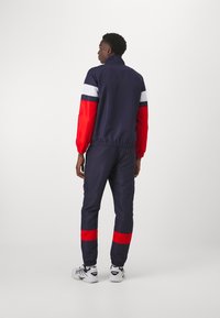 Navy track jacket with red sleeves, white stripes; matching pants with red accents. Fabric appears smooth, featuring elastic cuffs and waistband.