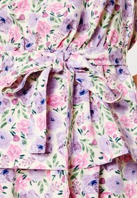 Vestido floral com um fundo bege claro e flores cor-de-rosa e roxas. Apresenta um cinto de tecido amarrado e uma camada de babados na barra.