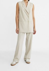Kobieta w beżowej, bezrękawnikowej, podwójnie zapinanej marynarce, pasujących szerokich spodniach i beżowych trampkach z jasnoniebieskimi detalami.