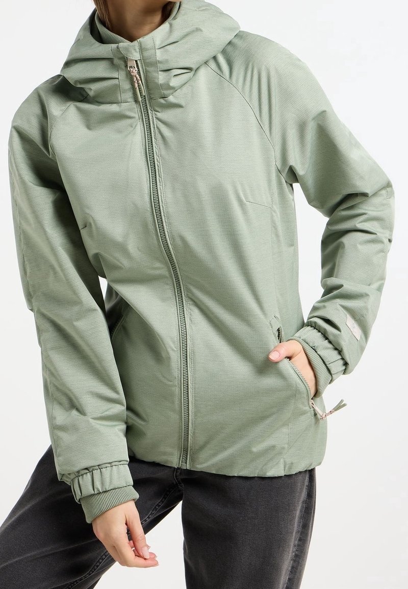 Personne portant une veste à capuche vert clair avec fermeture éclair devant et poches, une main dans la poche, associée à un pantalon foncé.