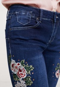 Mörkblå jeans i denim med blommig brodyr i rött och grönt, kompletterade med små metallstuds längs den nedre delen av benet.