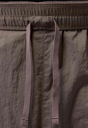 Gros plan de la ceinture d’un pantalon en tissu marron avec un élastique froncé et deux cordons plats pendants au centre.