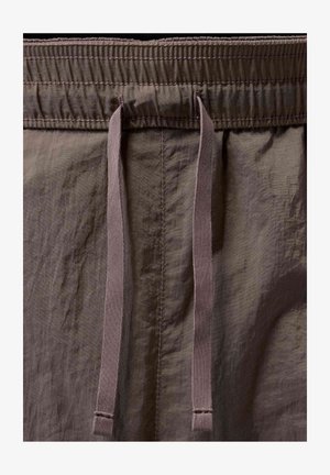 Gros plan de la ceinture d’un pantalon en tissu marron avec un élastique froncé et deux cordons plats pendants au centre.