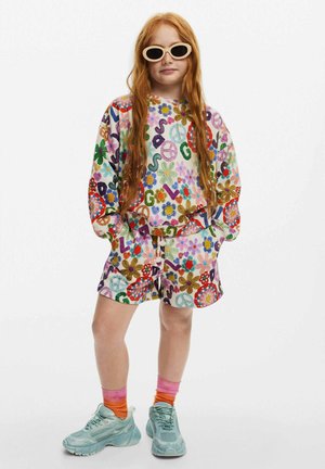 Conjunto de sudadera y pantalones cortos colorido y con patrones que presenta diseños florales, símbolos de paz y colores vibrantes. Combinado con zapatillas azul claro y gafas de sol.