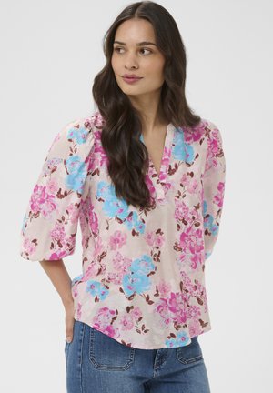 Žena s dlhými tmavými vlasmi oblečená v ružovej blúzke s modrými a fialovými kvetinovými vzormi a modrých džínsach, pozerajúca sa do strany.