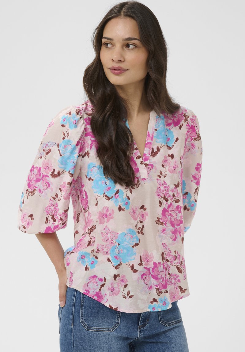 Femme aux longs cheveux foncés portant un chemisier rose avec des imprimés floraux bleus et violets et un jean bleu, regardant sur le côté.