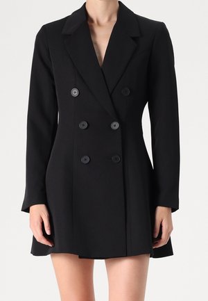Femme portant une robe blazer noire à double boutonnage avec six boutons, manches longues et col à revers en encoche.
