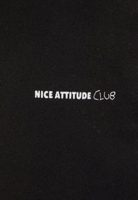 Tessuto nero con una superficie liscia, caratterizzato da testo ricamato bianco: "NICE ATTITUDE CLUB", con la parola "CLUB" in uno stile leggermente diverso.