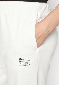Hand in der Tasche von weißen Lacoste-Sweatpants mit schwarzem Logo-Text und sichtbarem elastischem Bund.