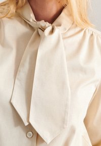 Beige blouse with a bow tie collar, button closure, soft texture, and slight sheen, featuring gathered details at the shoulders.
