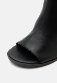 Gros plan d'une chaussure noire en cuir à bout ouvert avec une texture lisse et un design minimaliste sur fond blanc.