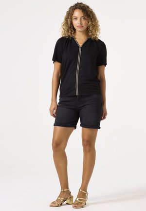 Zwarte top met korte mouwen, een kraag en een rits aan de voorkant, in combinatie met donkere denimshorts en goudkleurige sandalen met bandjes. Eenvoudig, casual ontwerp.