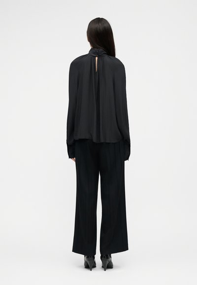 Femme aux longs cheveux bruns, vue de dos, portant un chemisier noir ample, un pantalon large noir et des chaussures à talons noires, sur un fond uni.