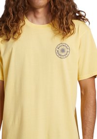 Camiseta de algodón amarilla con un logo redondo con diseño de sol en el lado izquierdo del pecho. Mangas cortas y un corte relajado. Textura suave.