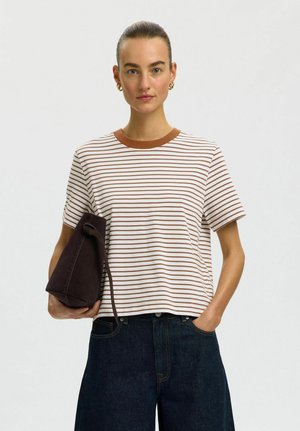 Donna con capelli raccolti indossa una maglietta a righe bianche e marroni, jeans scuri, orecchini a cerchio dorati, tiene una borsa marrone scuro.