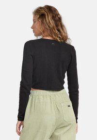 Black crop sweater with a ribbed texture and long sleeves. Notable details include a small logo at the back and a fitted design. Paired with light green corduroy pants.