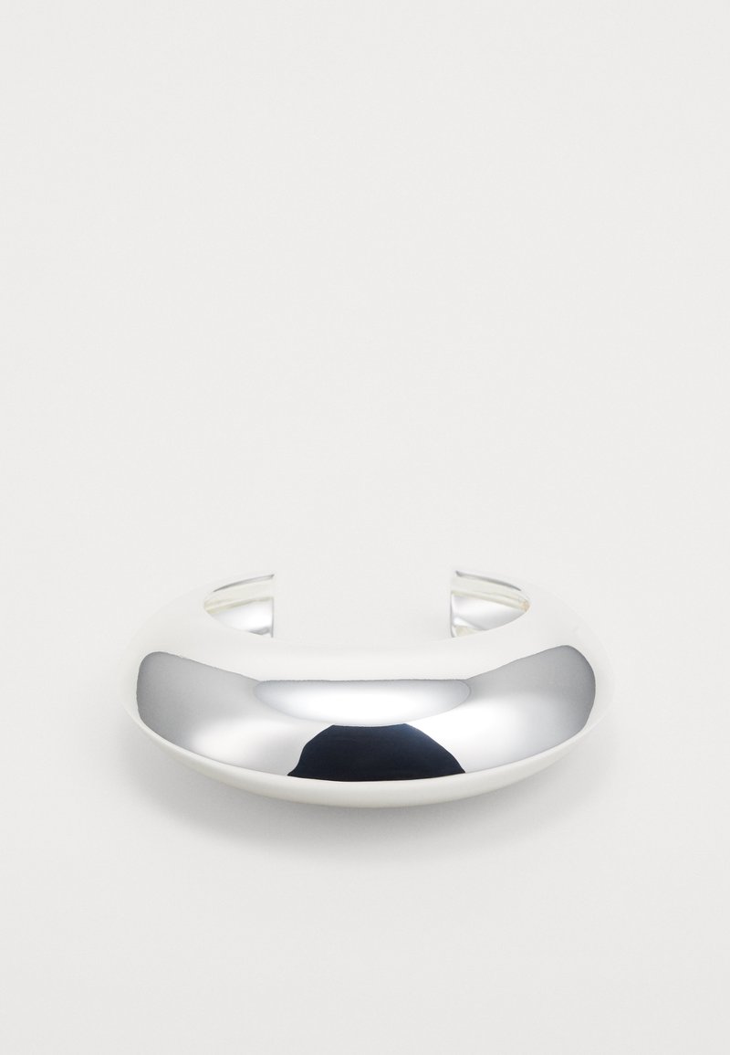 Brățară din metal argintiu, cu o suprafață netedă și reflectorizantă, capete deschise și un design minimalist, rotunjit.