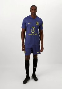 Męski piłkarz ubrany w ciemnoniebieski strój Atlético Madryt z żółtymi logo, czarne skarpetki i czarne korki, stojący na białym tle.