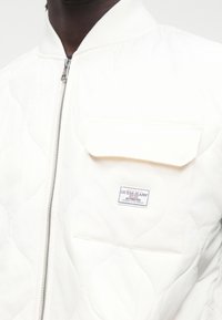 Artimas asmens, dėvinčio baltą siuvinėtą striukę su priekiu esančiu užtrauktuku ir kišene, pažymėta „Guess Jeans Authentic“.