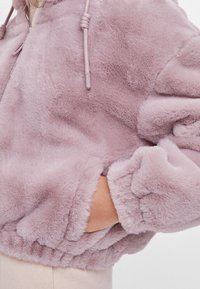 Zachte, puffy lavendel nep-vachtjas met elastische manchetten en hand in de voork pocket, gedragen over lichtbeige broek.