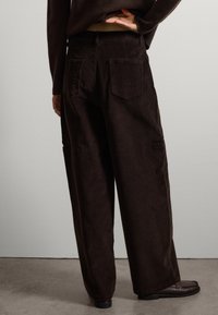 Pantaloni a gamba larga in corduroy marrone scuro con tasche posteriori e superficie testurizzata, abbinati a calzature scure. Il design mette in evidenza comfort e stile.