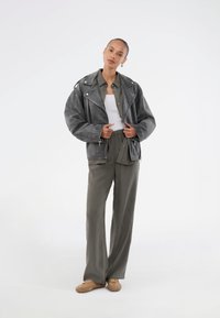 Veste de motard en cuir gris avec des zippers proéminents, assortie à un pantalon olive vert ample et des baskets beiges. Le mannequin se tient sur un fond neutre.