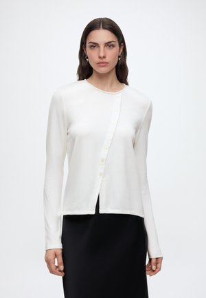 Femme aux longs cheveux foncés portant un cardigan blanc asymétrique boutonné et une jupe noire, debout devant un fond clair uni.