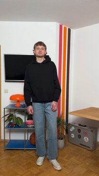 Mladý muž s tetovaním v čiernej mikine s kapucňou a modrých rifliach stojí v modernom priestore s farebnými pruhmi na stenách, rastlinami a kovovým kufríkom.