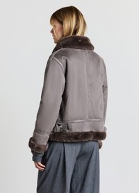 Veste en suède grise avec un col et des poignets en fourrure brune douce. Dispose d'une taille ceinturée et d'un tissu texturé. Modèle vu de dos.