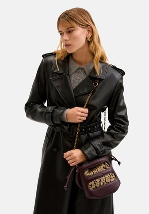 Sort faux læder trenchcoat med bælte i taljen, parret med en grå skjorte med krave og en lille lilla håndtaske med et mønstret forside.