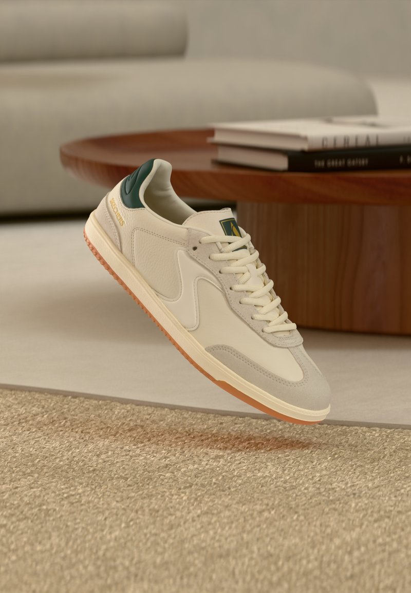 Μπέζ και λευκό αθλητικό παπούτσι με πράσινη λεπτομέρεια στη φτέρνα και καφέ σόλα να αιωρείται πάνω από ένα υφασμάτινο μπεζ χαλί, με ένα ξύλινο τραπέζι και στοιβαγμένα βιβλία στο φόντο.