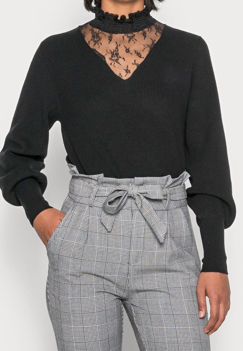 Femme portant un pull noir à manches longues avec un col en dentelle et un pantalon gris à carreaux taille haute, noué avec une ceinture en tissu.