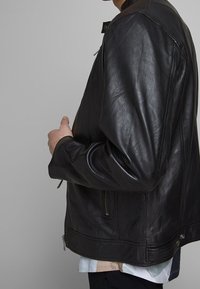 Zwarte leren jas met een gladde textuur, voorzien van een ritssluiting, zijzakken en een bruine kraagaccent. Iets open gedragen, waardoor een shirt eronder zichtbaar is.