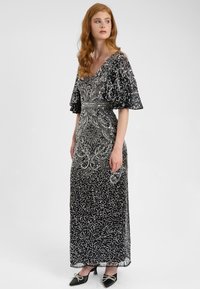 Abito da sera nero con intricati ricami in argento, maniche corte a volant e un corpetto aderente, che scende in una lunga gonna. Tacchi neri completano il look.