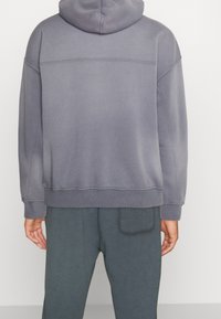 Henkilö, jolla on harmaa huppari ja tummanharmaat housut, kuvattuna takaa valkoista taustaa vasten.