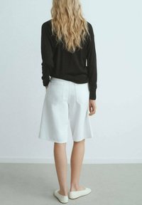 Shorts in denim bianco con vestibilità ampia, dotati di tasche laterali e tasche posteriori a toppa, abbinati a un top nero a maniche lunghe e scarpe bianche senza lacci.