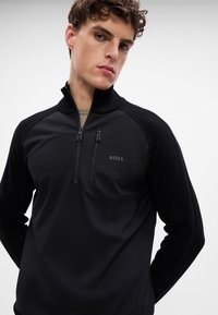 Črna zip-up pulover s stoječim ovratnikom, ima dva sprednja žepka na zadrgo, logo na prsih in teksturirane rokave. Splošna gladka tkanina.