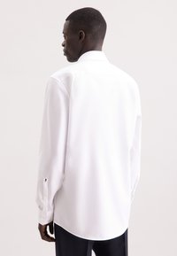 Camicia bianca a maniche lunghe con colletto classico e polsini abbottonati, realizzata in tessuto morbido con orlo dritto. Dettaglio di branding minimale visibile.