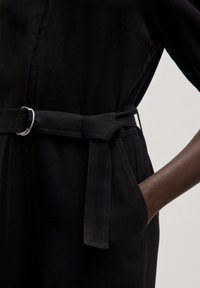 Zwart jumpsuit met een ceintuur in de taille, voorzien van een zilveren ringgesp, zijzakken en een zachte textuur. Geschikt voor verschillende gelegenheden.