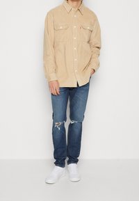 Beige manchester-skjorta med två bröstfickor, i kombination med slitna blå jeans och vita sneakers, stående mot en enfärgad bakgrund.