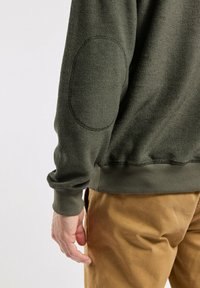 Sweat-shirt vert texturé avec des poignets côtelés, doté d'une patch en tissu ovale sur le coude. Porté sur un pantalon khaki à coupe décontractée.