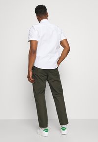 Witte overhemd met korte mouwen en een button-down kraag, gecombineerd met olijfgroene broek. Het model draagt witte sneakers met groene accenten.