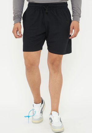 Uomo con pantaloncini elasticizzati neri in vita, maglia grigia a maniche lunghe e scarpe da ginnastica bianche con etichetta blu, in piedi su sfondo bianco.