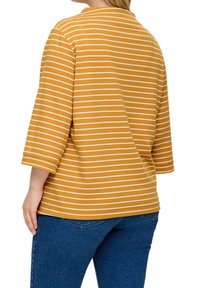 Mosterdgele top met witte horizontale strepen, met driekwartmouwen en een losse pasvorm. Zacht materiaal met een gladde textuur.