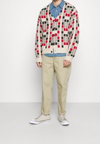 Stickad kofta i krämfärg med röda, svarta och grå geometriska mönster; lager på en denimskjorta; kombinerad med beige byxor och grå sneakers.