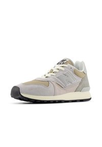 Sneaker in camoscio grigio chiaro e beige con sovrapposizioni testurizzate, lacci bianchi, linguetta imbottita e suola in gomma nera. Presenta un logo ben visibile.