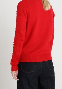 Pull rouge en maille avec des détails en relief en forme de boules sur les manches, poignets et ourlet côtelés, porté avec un jean en denim foncé.