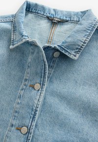 Chemise en denim bleu clair avec un col pointu, dotée de boutons en métal argenté et de détails de couture visibles. Le tissu est texturé et légèrement décoloré.